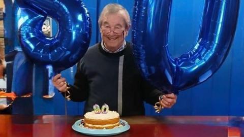Colin Morris holding two blue balloons that say 80 and standing in front of a birthday cake smiling at the camera
