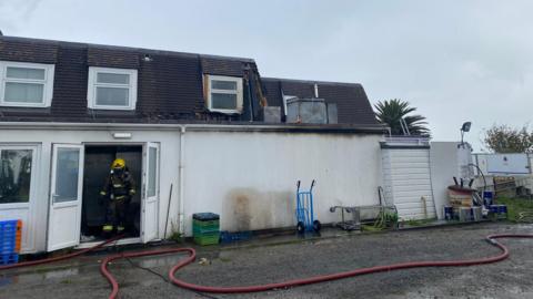 A fireman wearing breathing apparatus leaves the door of a fire-damaged hotel, with hoses across the yard.