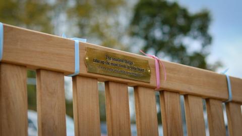 A memorial bench with blue and pink ribbons and a plaque honouring baby loss is outdoors beneath the blue sky.