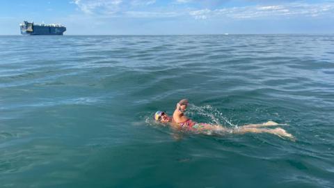 A woman swims in the sea wearing a brightly coloured swimming costume, goggles and a swimming cap. She is swimming front crawl with one arm out the water. In the distance is a large tanker ship and blue sky.