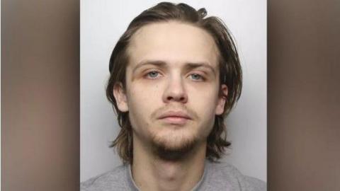 A police mugshot of Jack Naylor, who has long brown hair and a whispy brown goatee. He stares into the camera with a blank expression.