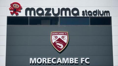 A view of the front of Morecambe's Mazuma Stadium completing with club badge and the words 'MORECAMBE FC' written