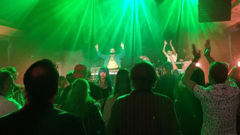 The stage is bathed in green light as Flamingods perform. The camera shows two men on stage, with their arms aloft encouraging the audience to clap along.
The camera is behind the audience and shows their back views, apart from one woman who has turned round.