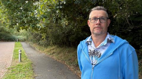 Paul Oakley with short dark hair and slight beard wearing a blue hoodie over a patterned T-shirt stands next to a tar-and-stone footpath with grass on either side, running past a hedge. There is a hedge running across the path in the background.