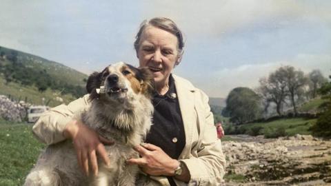 A woman with grey hair on a moor wearing a mac and a black cardigan hugs a white border collie dog.