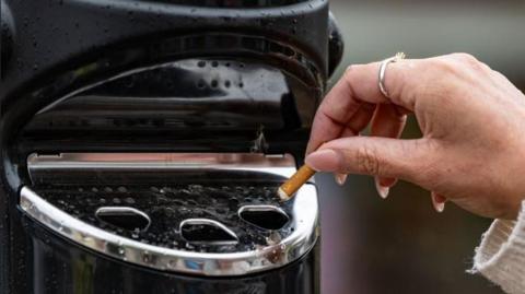 A hand holding a cigarette butt over a black and silver bin.