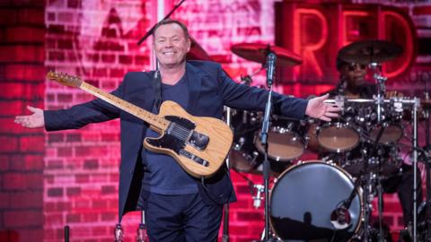 Ali Campbell, on stage with his arms outstretched, is wearing a black suit with a dark-coloured T-shirt. There is a drum kit and a drummer behind him, and a backdrop showing red-lit bricks with the word 'red' in capital letters in the appearance of a neon sign.
