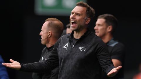 Derby County manager John Eustace opens mouth wide and shouts whilst holding his hands out wide. He wears a black Derby County quarter zip top