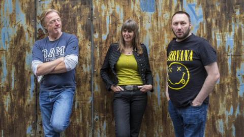 Two men and a woman are standing in front of a wall that is metal and rusted. The man on the left has short blonde hair and a white goatee. The woman has brown shoulder length hair. The man on the right has short black hair with black facial hair. He is wearing a black and yellow Nirvana t-shirt