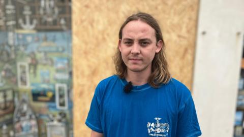 A man with long hair wearing a blue t-shirt stood in front of a boarded-up window.