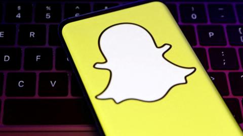 A smartphone showing Snapchat's logo, a white, faceless ghost figure on a yellow background, in front of a keybaord.