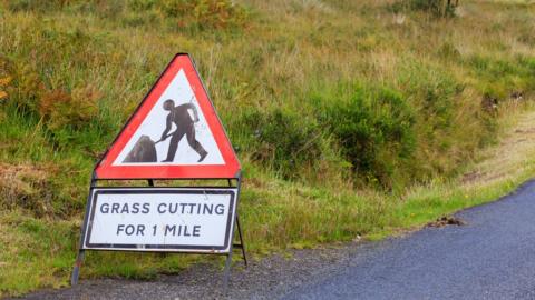 UK Triangle road sign warning of grass cutting for one mile