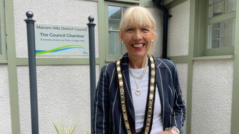 Beverley Nielsen pictured beside a sign for Malvern Hills District Council. She has blonde hair which has been tied back and is wearing a navy blue jacket with white stripes. She is also wearing gold jewellery and a ceremonial gold chain around her neck.