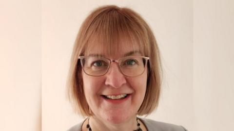 Headshot of Jo Walker, a middle-aged woman with a strawberry blonde, fringed bob and light rimmed glasses, smiles. She is wearing a black beaded necklace and the shoulders of her grey top can be seen. She has blue eyes and is standing against a plain, cream-coloured background.