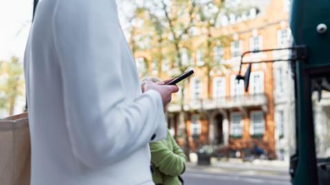 Woman in white cardigan looks at mobile phone in a London street