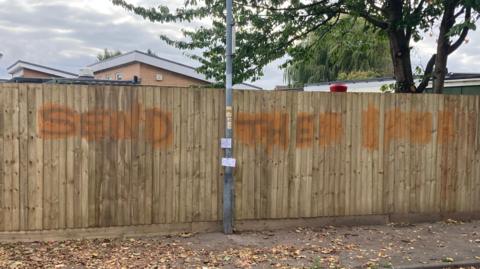 The words "send them home" daubed on a fence panel in Boston. There is a tree and a building in the background