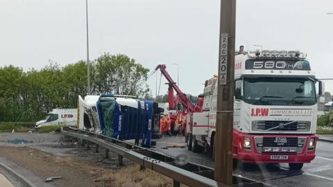 The image shows the aftermath of a road incident on a dual carriageway. In the mid-ground, a large blue lorry is lying on its side across the edge of the road, partially resting against a metal crash barrier. Several recovery vehicles, including a red heavy-duty tow truck and another lorry marked “J.P.W Commercials Ltd,” are positioned nearby, with workers in high-visibility clothing gathered around the overturned vehicle.
