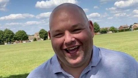 Gary Stanley, standing outside, by a field, smiling. He is wearing a blue top. Buildings and trees are behind him in the field. 