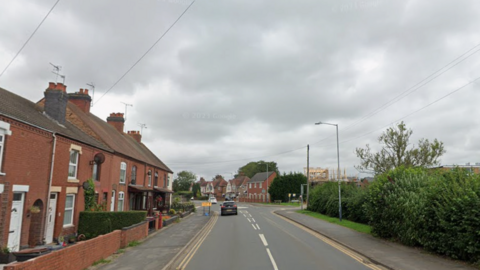 A residential street with houses on one side, bushes on the other and a car driving along the road.

