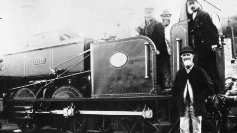 A black and white photograph of a steam locomotive with characters who used to run it on the footplate and a bearded gentleman wearing a bowler hat standing in front of the train