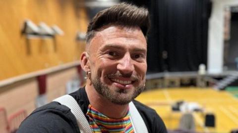 A smiling Jake Quickenden looking straight at the camera. He is wearing a multi-coloured striped top with black sleeves and is sitting backwards on a theatre seat.