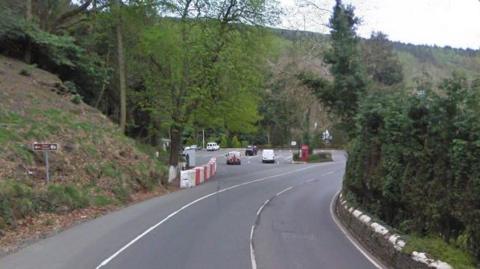 Part of the TT course approaching the bend at Glen Helen. The road is lined with green trees an embankment and a low stone wall painted white on the top. Some cars are parked in the car park at Glen Helen.