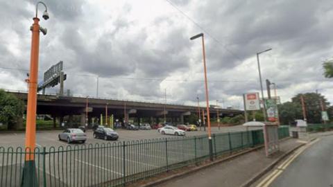 A Google Maps screenshot of a train station car park. There is a bus stop next to the car park, which has a sign bearing a red and white railway symbol. There are orange lampposts lining the edge of the car park. 