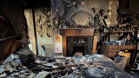 A room badly damaged by fire has ash and rubble strewn across the floor. A fireplace and desk on the far wall seem mostly undamaged. The walls are blackened with ash.