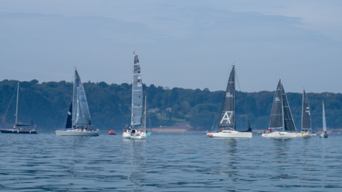 Eight sailing boats on the waters, grey and blue sails, clear blue skies behind with greenland in the background