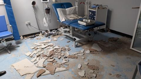 A reclining patient chair with a pillow is seen in a treatment room, with rubble and cement scattered across the floor and on surrounding equipment. 