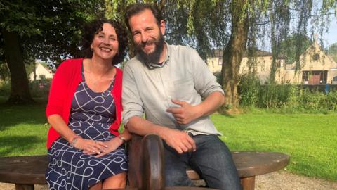 Amanda Woodhead and Dom from the Repair Shop sat on the refurbished aircraft bench. They are both smiling into the camera.