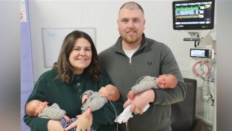 A woman and a man stand next to each other on a hospital ward. The woman is holding two newborn babies and the man is holding one. The babies are in matching knitted jumpers.  