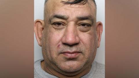 A police mugshot of a man with short black hair wearing a grey prison top