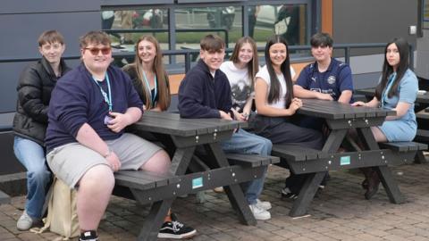 A group of male and female students, all aged 17, sit together, smiling, at two picnic tables in front of their college building