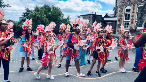 Children are wearing red costumes and dancing within a school's ground.