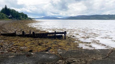 A muddy area leads down a gentle slop into a loch with mountains in the background on a cloudy day