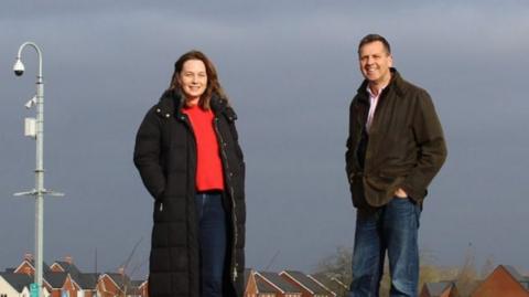 A woman and a man are stood near a pole and a CCTV camera is visible nearby.