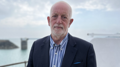 Steve Luce pictured with the sea visible behind him. He's wearing a navy blazer and a blue and white pinstripe shirt. He has short white buzzed hair and beard. 