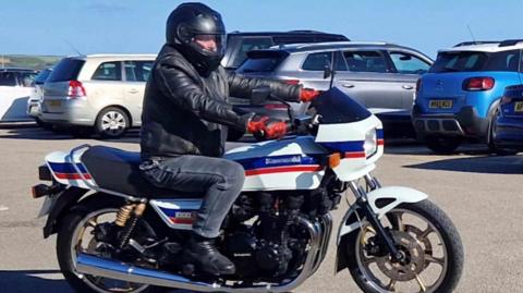 David Staveley, wearing a black leather motorcycle jacket, black crash helmet and black jeans, riding a white Kawasaki motorbike with blue and red stripes in a car park