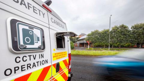 A police safety camera van parked by the side of the road. A hatch at the back of the van is open. On the rear door of the van it says CCTV in operation. A blurry blue car can be seen passing the van.