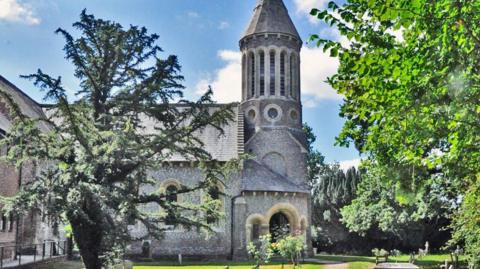 A lovely sunny day with a church-looking type building behind a large tree with more trees on the right-hand side