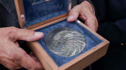 The Suffolk Medal in a wooden box, it is silver with a wave engraved on it. It's being held in a blue fabric frame.