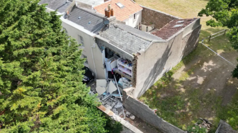 A picture of a house that was damaged in an explosion. There is a property with part of the back wall missing across two storeys and plaster spread everywhere.
