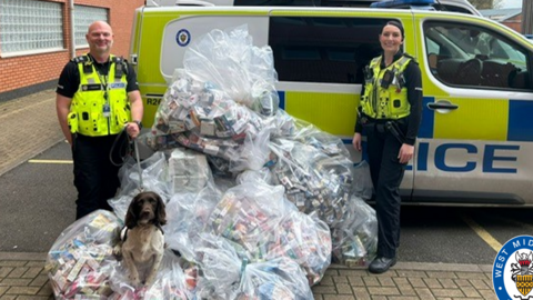 Police officers pictured wearing police uniform in front of a police van. The image includes plastic bags piled high and full of seized products, with a sniffer dog sat on the top of one of the bags. 