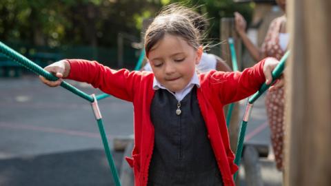 A child walks along a climbing frame, gripping two green ropes for balance. She is wearing a bright red cardigan over a dark grey pinafore dress with a white shirt underneath. Hair brown is tied back in a ponytail. The school playground is blurred in the background.