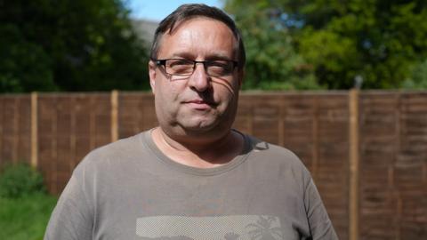 Mark Sumner looking into the camera with a neutral expression standing in a garden with a wooden panelled fence behind him with grass, trees and blue sky visible
