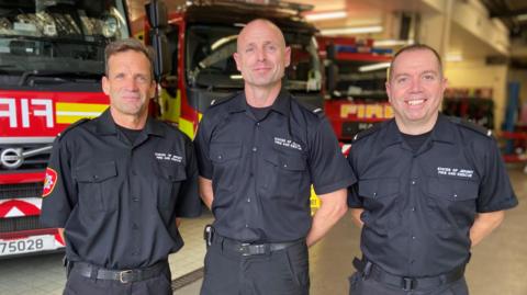 Firefighters Nick Willis, Chris Sweeny and Jonny Burch face the camera wearing dark uniform with a logo saying States of Jersey Fire and Rescue with fire engines behind.