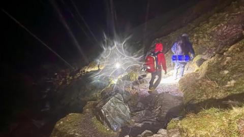 Two mountain rescuers with torches climbing a mountain in the dark. One is wearing a red jacket, black trousers and a red bag. The other is wearing a blue coat and a blue bag. They are walking along a rocky path.
