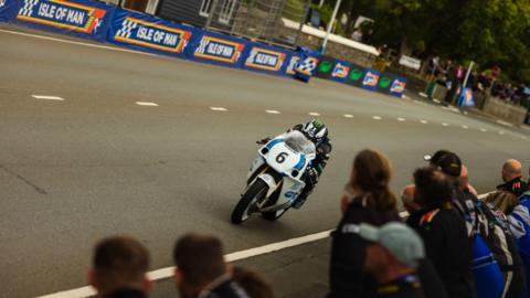 Michael Dunlop, wearing mainly black leathers and a black-and-white helmet with a bright green logo, rides his motorbike on Glencrutchery Road with spectators looking on.