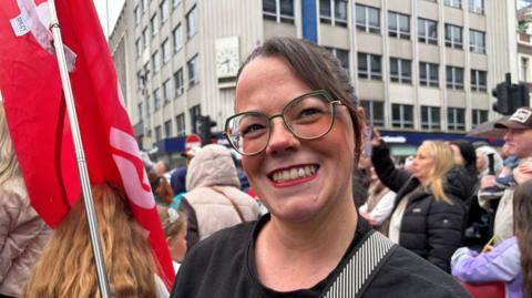 A woman with long brown hair and clear-lensed glasses wears red lipstick as she smiles to the camera. She holds a red flag in her left hand. Behind her his a large crowd of people, out-of-focus. 